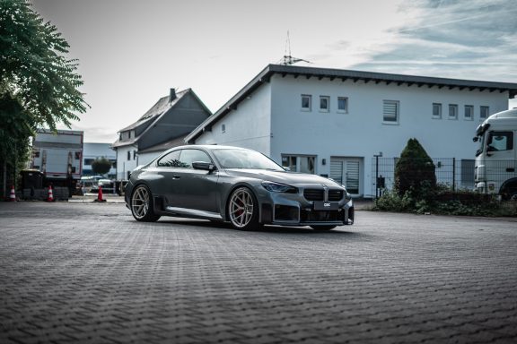BMW M2 G87 mit Edelweiss-Rädern und Carbon-Frontdetails in einer tiefen Dreiviertelansicht vor einem Gebäude.